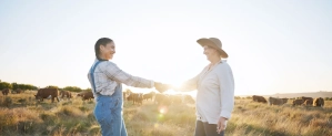 two woman holding hand with a cow herd in background